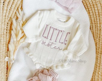May include: A cream-colored baby outfit set with the embroidered text "LITTLE sister". The set includes a long-sleeved romper, a headband, and booties, all in a soft, knitted design. The outfit is displayed on a white blanket inside a woven basket.