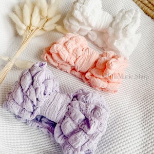 May include: Three fabric hair bows in different colours: white, pink, and purple. The bows are made of a textured fabric and are arranged on a white surface.