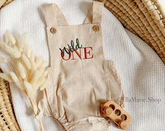 May include: A beige linen baby romper with adjustable straps and a red embroidered design that says "Wild One".