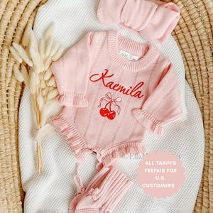 May include: A light pink baby outfit set, including a romper, headband, and booties. The romper features the name "Kaemila" embroidered in red, along with a cherry design. The set is made of a soft knit fabric and is displayed on a white blanket.