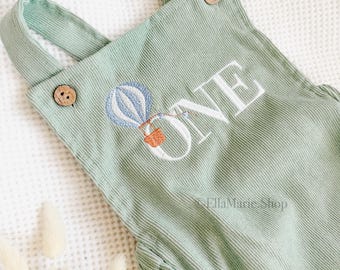 Könnte beinhalten: Ein salbeigrüner Cord-Overall mit zwei braunen Holzknöpfen an den Trägern. Die Vorderseite ist mit einer blau-weißen Heißluftballon-Grafik und dem weißen Schriftzug ONE bestickt. Das Kleidungsstück hat eine texturierte, gerippte Stoffoberfläche.