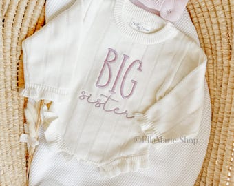 May include: Cream-colored baby sweater with the embroidered text "BIG sister" in lavender. The sweater has a ruffled hem and sleeves, and is paired with a matching pink bow. The items are displayed in a woven basket.