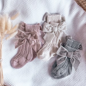 May include: Three pairs of baby socks with bows. The socks are pink, beige, and grey. The bows are made of velvet and have a sparkly finish.