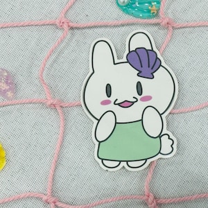 May include: A white bunny with a purple seashell on its head is wearing a green dress. The bunny is smiling and has pink cheeks. The bunny is on a pink and white background.