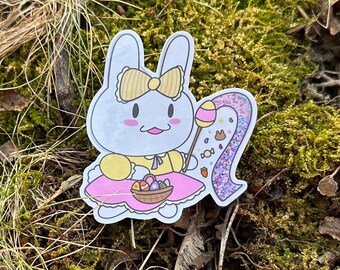 May include: A sticker featuring a cartoon bunny with a yellow bow, dress, and holding a wand. The bunny is holding a basket of Easter eggs and a glittery number two. The sticker is set against a mossy background.
