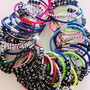 Op de afbeelding: Een verzameling kleurrijke elastieken armbandjes met woorden die zijn gespeld met zwarte letterkraaltjes. De woorden omvatten "Fearless", "True", "Rise", "Passion", "Dig", "Code", "You", "Believe", "Strong", "No", "Committ", "Sister", "Hight", "Pass", "One", "20", "T", "S", "B", "Shi", "30", "7", "00", "SS", "H", "R", "TRUEL", "<RONG", "G", "ENUN", "RISE", "EL", "EPASSI", "DEYOURSELF", "PASSIONA", "DIGODE", "F", "TRUEG", "1", "EP", "P", "CA", "CAN", "S", "S", "ar", "NOLM", "a", "378R", "51", "NEBR", "D", "M", "9120", "NOSTEN".