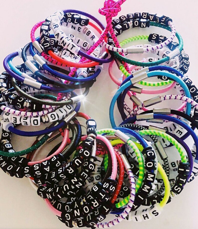 Op de afbeelding: Een verzameling kleurrijke elastieken armbandjes met woorden die zijn gespeld met zwarte letterkraaltjes. De woorden omvatten "Fearless", "True", "Rise", "Passion", "Dig", "Code", "You", "Believe", "Strong", "No", "Committ", "Sister", "Hight", "Pass", "One", "20", "T", "S", "B", "Shi", "30", "7", "00", "SS", "H", "R", "TRUEL", "<RONG", "G", "ENUN", "RISE", "EL", "EPASSI", "DEYOURSELF", "PASSIONA", "DIGODE", "F", "TRUEG", "1", "EP", "P", "CA", "CAN", "S", "S", "ar", "NOLM", "a", "378R", "51", "NEBR", "D", "M", "9120", "NOSTEN".