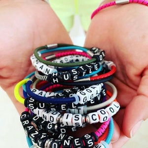Op de afbeelding: Een stapel kleurrijke armbanden met woorden die zijn gespeld in zwarte en witte kralen. De woorden zijn "Hustle", "Patience", "Success", "Goals", "Crazy", "Kindness", "Believe", "Cool", "Strong" en "Disco".
