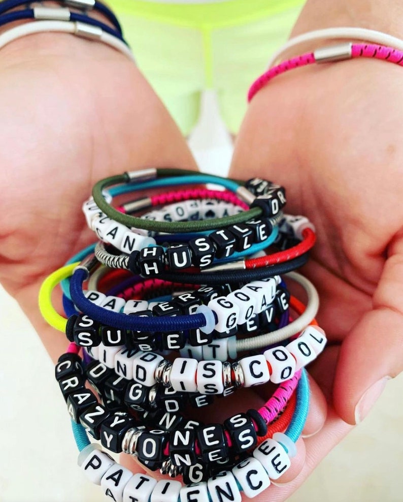 Op de afbeelding: Een stapel kleurrijke armbanden met woorden die zijn gespeld in zwarte en witte kralen. De woorden zijn "Hustle", "Patience", "Success", "Goals", "Crazy", "Kindness", "Believe", "Cool", "Strong" en "Disco".