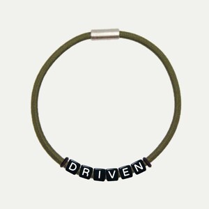 Op de afbeelding: Een groene elastische armband met een zilveren sluiting en zwarte letter kralen die het woord "DRIVEN" spellen.