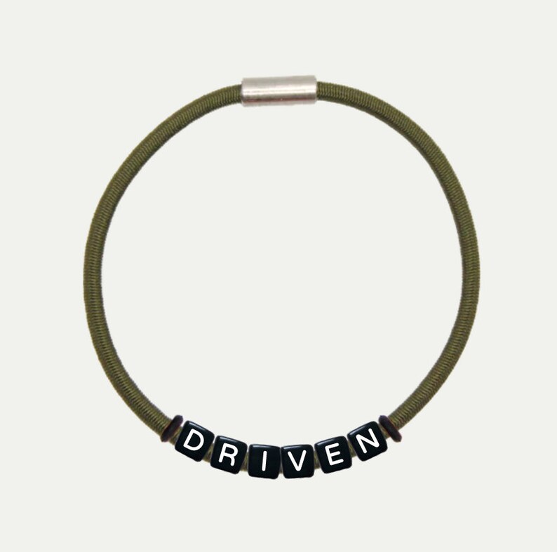 Op de afbeelding: Een groene elastische armband met een zilveren sluiting en zwarte letter kralen die het woord "DRIVEN" spellen.