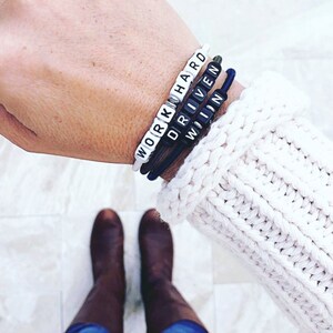Op de afbeelding: Een pols met drie armbanden met de woorden "WORK HARD", "DRIVEN" en "WIN" gespeld in witte letters op zwarte kralen.