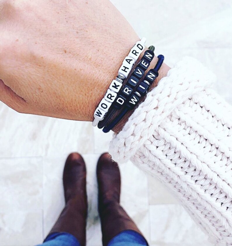 Op de afbeelding: Een pols met drie armbanden met de woorden "WORK HARD", "DRIVEN" en "WIN" gespeld in witte letters op zwarte kralen.