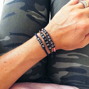 Op de afbeelding: Drie kralenarmbanden met de woorden "WIN", "DRIVEN" en "FORWARD" gespeld in zwarte kralen op een roze, groene en blauwe elastiek.