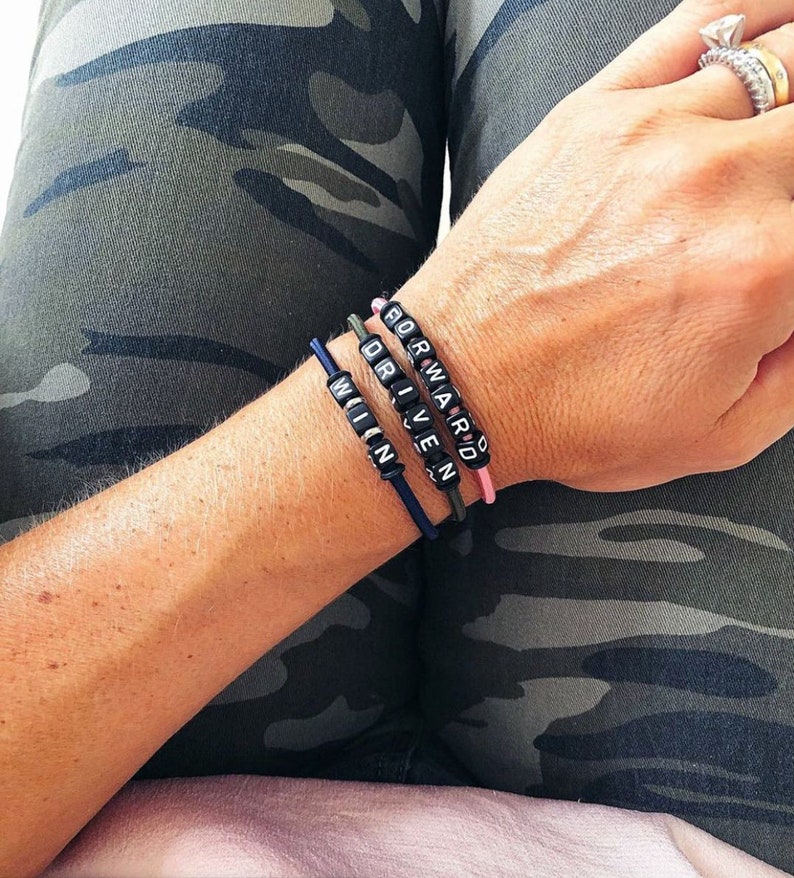 Op de afbeelding: Drie kralenarmbanden met de woorden "WIN", "DRIVEN" en "FORWARD" gespeld in zwarte kralen op een roze, groene en blauwe elastiek.