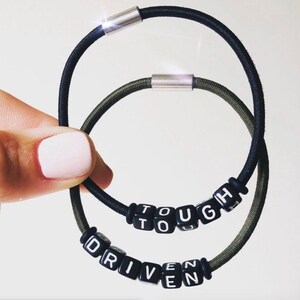 Op de afbeelding: Twee elastische armbanden met zilveren metalen sluitingen. Een armband is zwart en de andere is groen. Beide armbanden hebben zwarte letter kralen die de woorden "TOUGH" en "DRIVEN" spellen.