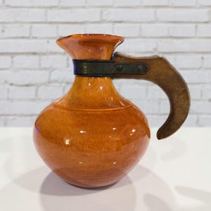 Może przedstawiać: Błyszczący, pomarańczowo-brązowy ceramiczny dzbanek z drewnianym uchwytem i ciemną metalową opaską. Dzbanek ma zaokrąglony korpus i rozszerzoną krawędź. Uchwyt jest zakrzywiony i przymocowany metalowym osprzętem. Dzbanek stoi na białej powierzchni.