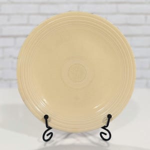 May include: A cream-colored ceramic plate with concentric rings and a central design, displayed on a black metal stand. The plate has a glossy finish and is set against a white brick wall background.