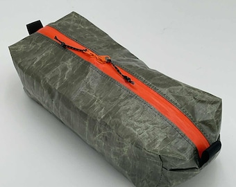 Peut inclure: Une trousse rectangulaire fabriquée dans un matériau translucide gris olive froissé. Elle est dotée d'une fermeture éclair orange vif sur le dessus, équipée de deux tirettes en cordon noir. Des boucles en tissu noir sont fixées aux extrémités.