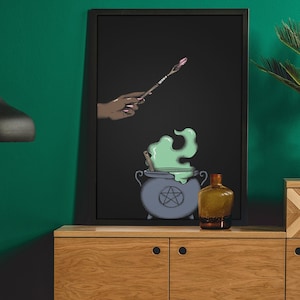 Puede incluir: Una impresión enmarcada en negro con una mano sosteniendo una varita mágica sobre un caldero con humo verde. El caldero tiene un pentagrama en el lateral. La impresión está en un estante de madera con una botella negra y una planta en maceta.