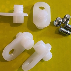 Könnte beinhalten: Eine Sammlung weißer Kunststoff-Möbelbeschlagteile, darunter mehrere Einschraubstifte, ein Stift mit flachem Sockel und mehrere kleine silberne Schrauben. Die Artikel sind auf einer gelb-grünen Oberfläche angeordnet.