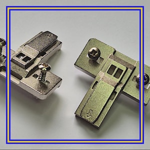 May include: Two metal cabinet hinges with screws. The hinges are silver and have a rectangular shape with various cutouts and a small arrow. The screws are silver and have a star-shaped head.