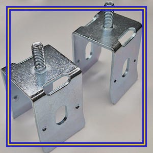 May include: Two galvanized steel corner brackets with threaded bolts. Each bracket has a U-shape with rounded edges and several holes for mounting. The bolts are centered on the top of each bracket.