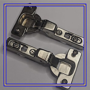 May include: Two silver-colored cabinet hinges with multiple screw holes and a pivoting joint. The hinges are made of metal and are designed for mounting on cabinet doors. The brand name "LAMA" is visible on one of the hinges.