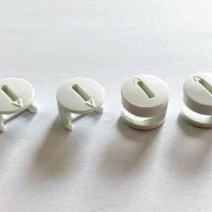 May include: Four white plastic circular fasteners arranged in a row. Each fastener features a flat top surface with a central rectangular slot and a small embossed arrow pointing toward the slot. The components have a side opening design.
