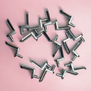 Op de afbeelding: Een close-up van een groep zilveren metalen plankbeugels op een roze achtergrond. De beugels hebben allemaal dezelfde vorm, met een rechte hoek en een kleine pin aan het uiteinde.