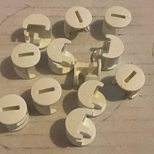 Könnte beinhalten: Eine Gruppe von 11 cremefarbenen Plastikkappen mit einem Schlitz in der Mitte. Die Kappen sind in einem Kreis auf einer hellbraunen Oberfläche angeordnet.