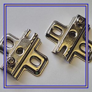 Pode incluir: Duas dobradiças de armário de metal prateado brilhante com fixadores de parafuso. As dobradiças têm uma forma retangular com bordas curvas e vários orifícios para montagem. O hardware é projetado para construção e reparo de móveis.