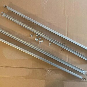 Op de afbeelding: Twee zilveren metalen plankbeugels met schroefgaten en zes zilveren schroeven op een bruin oppervlak.
