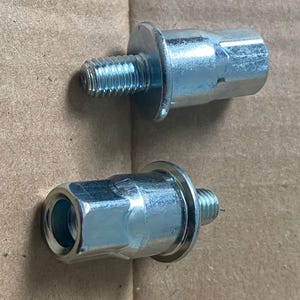 May include: Two silver-colored bolts with hexagonal heads and threaded shafts. The bolts have a circular flange near the head. The bolts are on a brown surface.