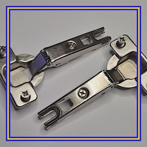 May include: Close-up of several silver cabinet hinges. Each hinge has a curved, circular base with a rectangular arm and a small screw. The hinges are made of metal and have a shiny finish. The background is a light grey.
