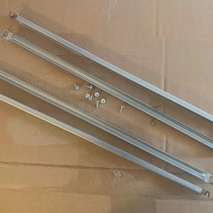 May include: Four silver metal shelf supports and several screws are arranged on a cardboard surface. The shelf supports are rectangular with a series of parallel ridges and mounting holes at each end. The screws are small and silver.