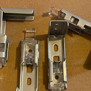 May include: A collection of silver-colored blind brackets and mounting hardware. The set includes L-shaped corner brackets and vertical brackets with clear plastic components. The hardware is designed for window treatments and home improvement projects.
