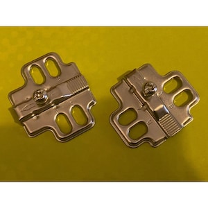 May include: Two silver metal cabinet hinges with screw fixings. Each hinge has a rectangular shape with rounded cutouts and a textured section. The hinges are displayed on a bright yellow background.