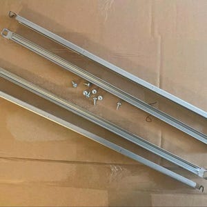 Op de afbeelding: Twee zilveren metalen beugels met schroefgaten en vier zilveren schroeven. De beugels liggen op een bruin oppervlak.