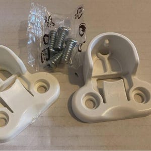 Może przedstawiać: Dwa białe plastikowe uchwyty do karniszy z otworami na śruby i mała torebka srebrnych śrub. Uchwyty mają zakrzywioną górę i płaską podstawę z dwoma otworami. Przedmioty znajdują się na jasnej powierzchni.