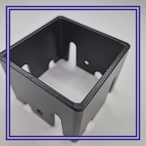 May include: A black, square-shaped metal object with cut-out sections on the sides. The object has a slightly textured surface and appears to be a component or part of a larger structure. The edges are defined and the overall shape is geometric.