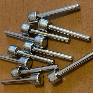May include: A collection of shiny, silver-colored metal pins with rounded heads. The pins are scattered on a brown surface, with their cylindrical shafts and domed heads clearly visible. The pins appear to be new.