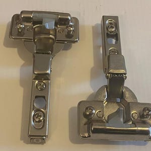 Peut inclure: Deux charnières de meuble argentées. Chaque charnière a une base rectangulaire avec des trous de vis et un bras pivotant. Les charnières sont conçues pour être montées sur une porte et un cadre de meuble.