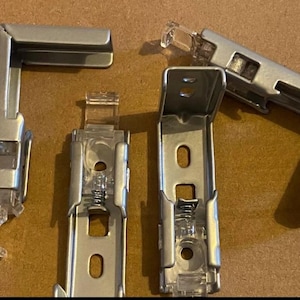 May include: Four silver-colored blind brackets are displayed against a brown background. Two are L-shaped, and two are rectangular with a spring mechanism. The brackets have clear plastic components and are designed for window treatments.