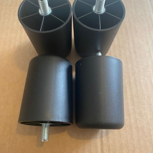 Könnte beinhalten: Vier schwarze Möbelbeine aus Kunststoff mit Gewindeeinsätzen aus Metall. Die Beine sind zylindrisch und haben eine glatte Oberfläche.