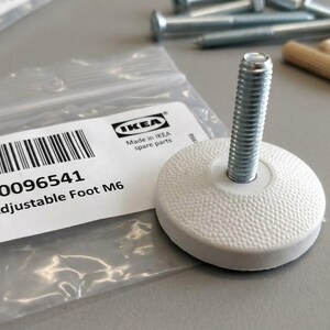 May include: An IKEA adjustable foot M6 with a white circular textured base and a silver metal threaded bolt. The part is accompanied by a clear plastic bag featuring a white label with a barcode, the text 10096541, and Adjustable Foot M6.