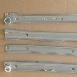 Può includere: Quattro guide per cassetti in metallo grigio chiaro, probabilmente per il montaggio di mobili. Le guide hanno vari fori e una rotella a un'estremità. Una guida è contrassegnata con "IKEA C/ 72419". Lo sfondo è marrone chiaro.