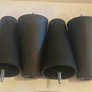 以下が含まれることがあります： 黒色の円錐形の家具の脚が4つ。取り付け用のねじ付き金属スタッドが付いています。脚は無地のマットな素材でできており、等間隔に配置されています。脚の高さは約15cmです。