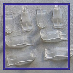 May include: A collection of clear plastic bottle-shaped components, some upright and some lying down. Each piece has a rounded top and a rectangular body. The components are arranged on a textured, light gray surface.