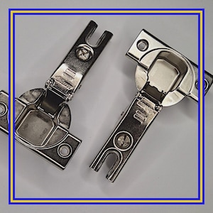 Può includere: Due cerniere per mobili argentate e lucenti sono mostrate su uno sfondo bianco. Ogni cerniera è aperta, rivelando i meccanismi interni e i dettagli delle viti. Le cerniere sono in metallo e sono progettate per mobili.